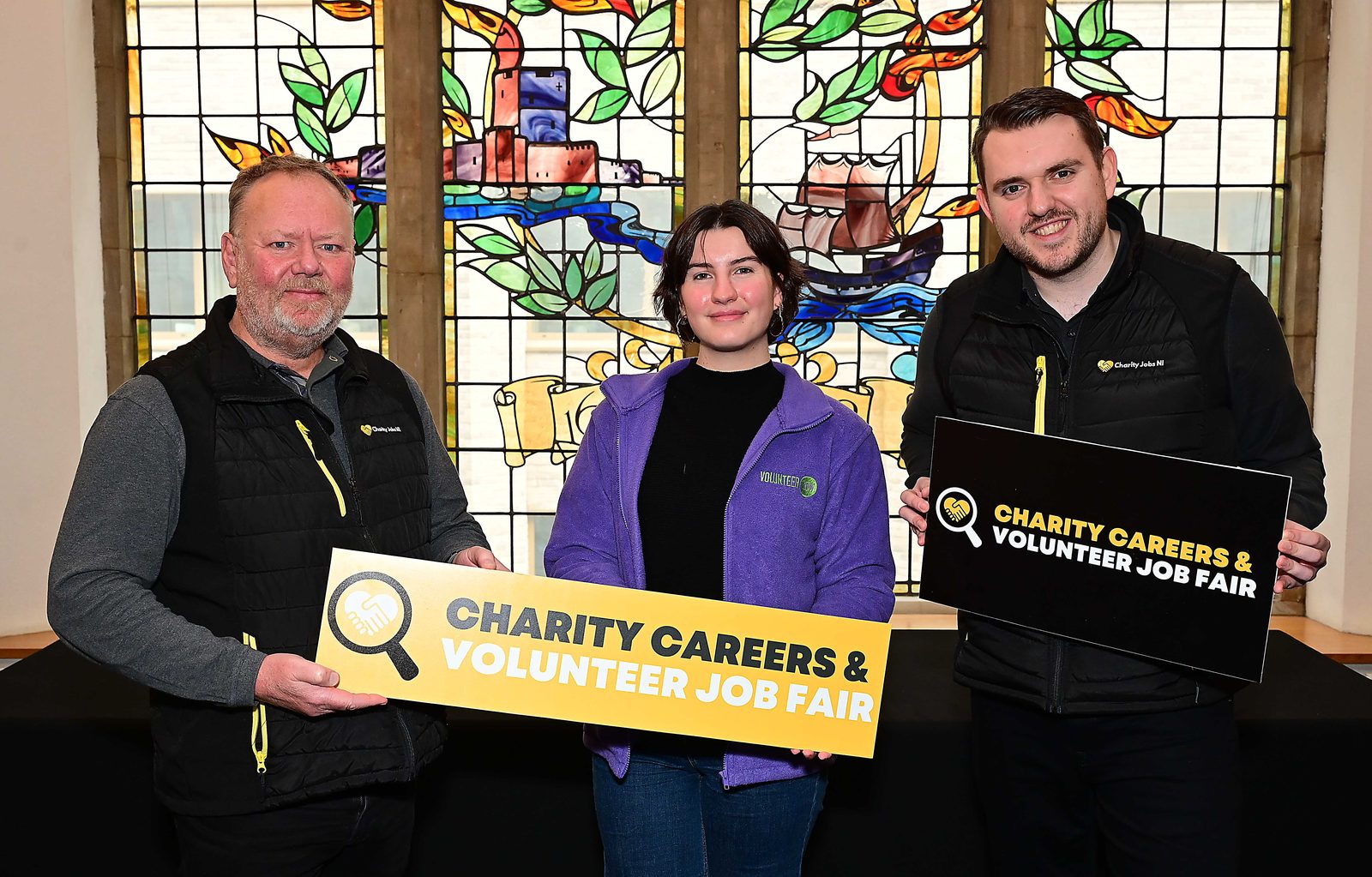 Three people holding Charity Careers & Volunteer Job Fair signs beneath a stained-glass window at Assembly Buildings, Belfast