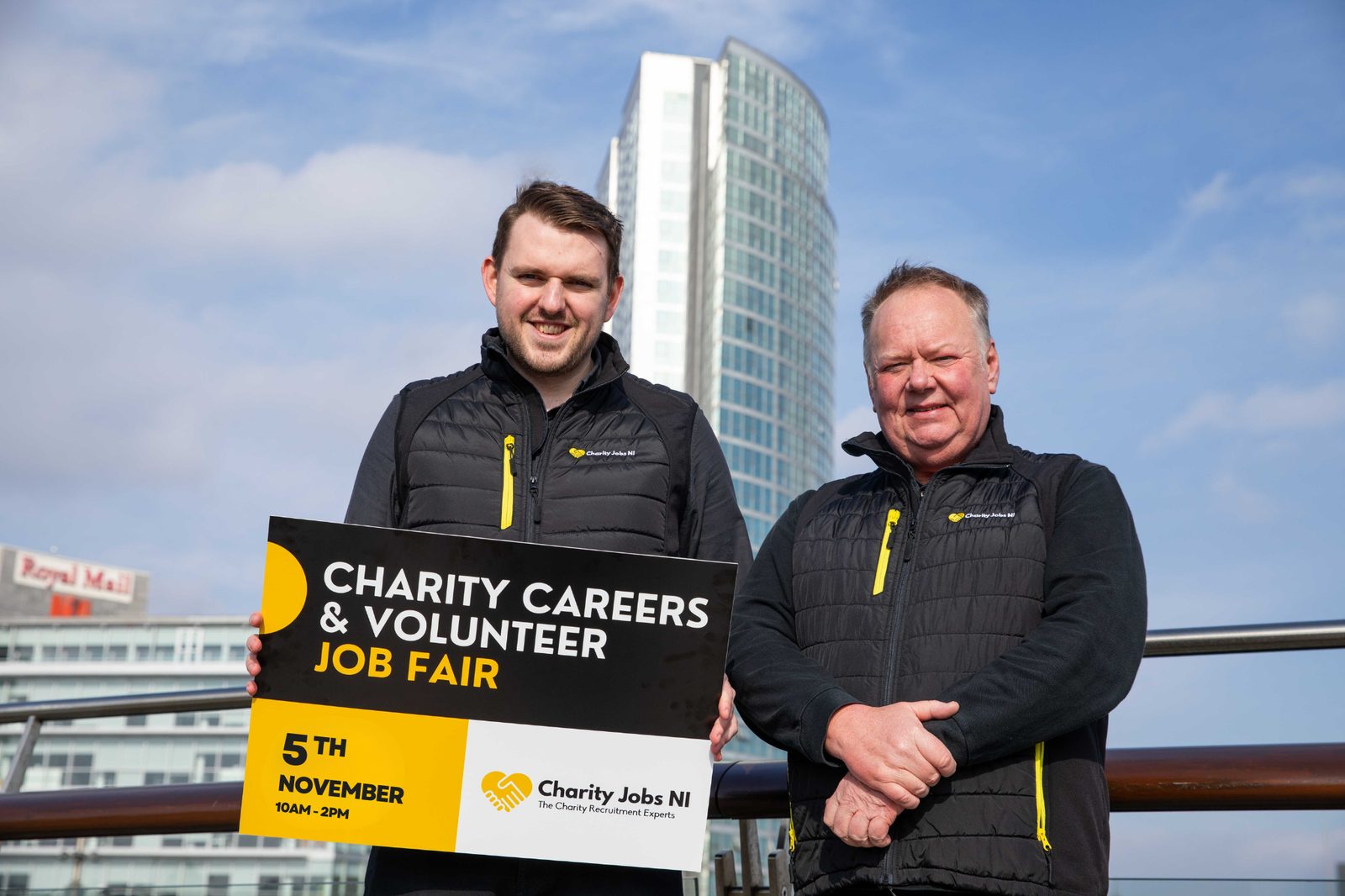 Two Charity Jobs NI representatives holding a Charity Careers & Volunteer Job Fair board with the Belfast skyline behind them