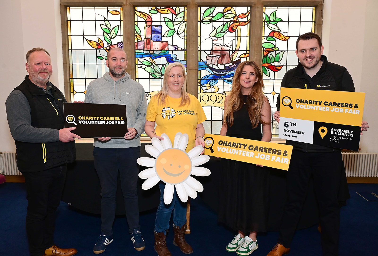 Five charity staff, one holding a large daisy mascot prop, with Charity Careers & Volunteer Job Fair boards at Assembly Buildings, Belfast