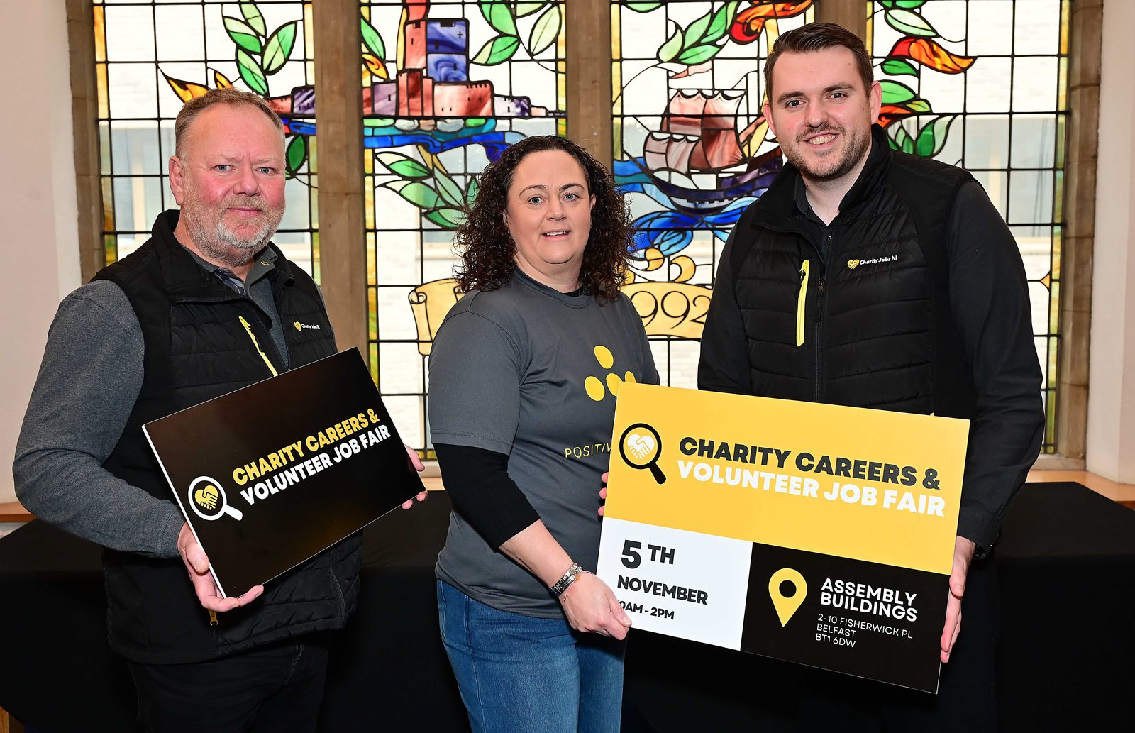 Three charity representatives holding Charity Careers & Volunteer Job Fair boards in front of a stained-glass window at Assembly Buildings, Belfast