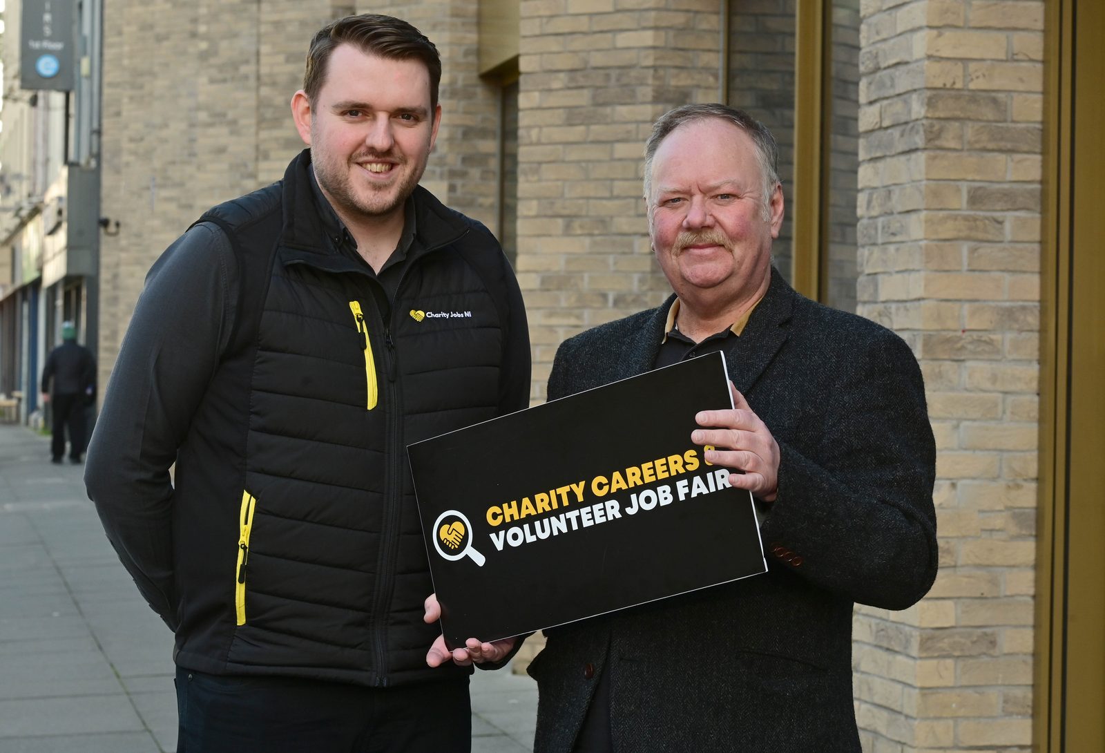 Two men holding a Charity Careers & Volunteer Job Fair board outside Assembly Buildings, Belfast