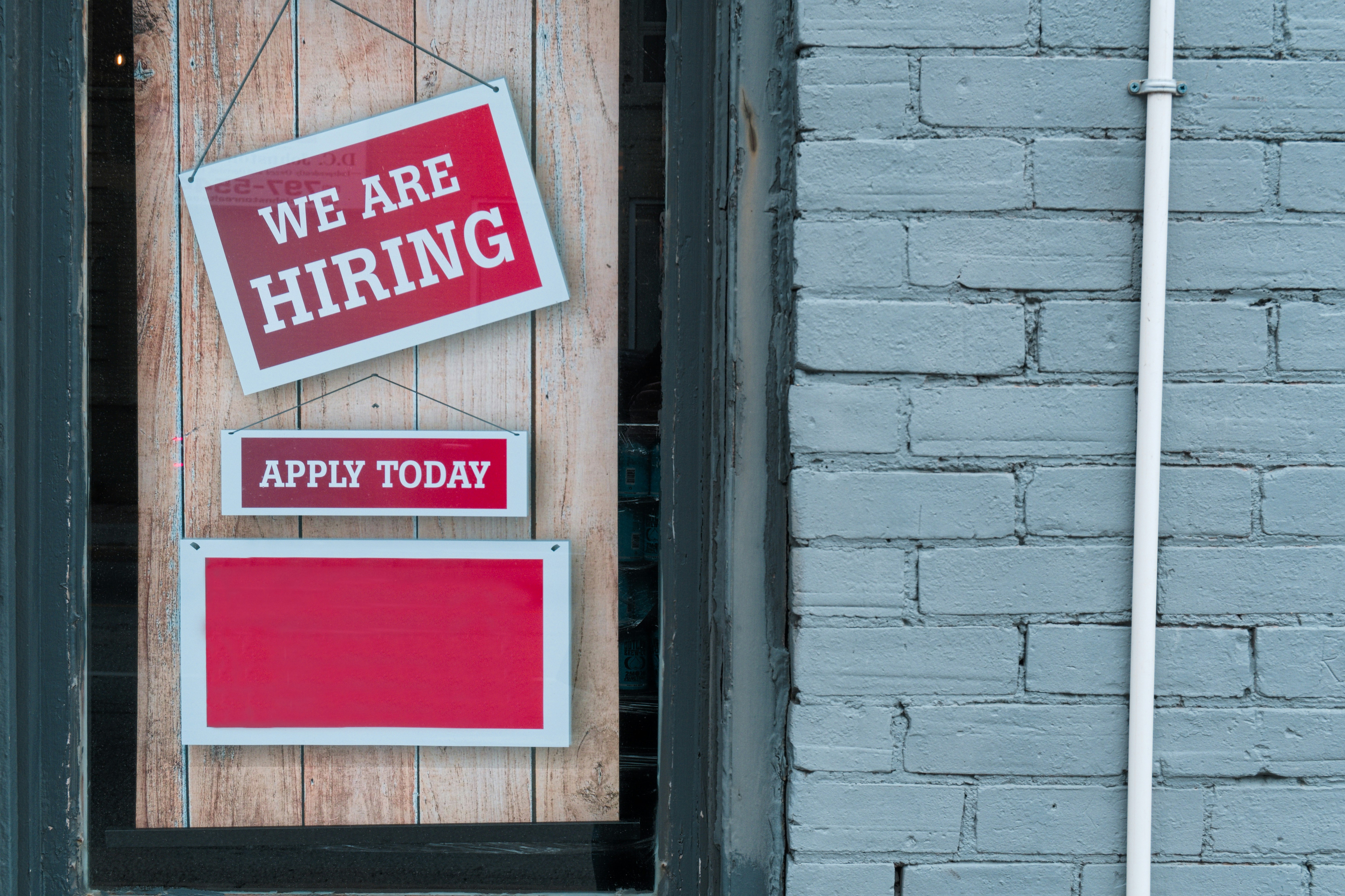 A red "We are hiring - apply today" sign hanging beside a grey painted doorway